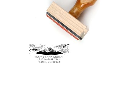 A light wood block stamp turned on its side next to a stamped image featuring a mountain and an address for Rory and Emma William