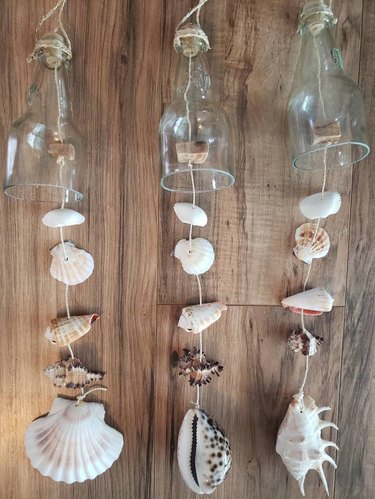 A trio of wind chimes made with half a clear glass bottle, seashells attached to twine and driftwood.