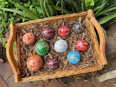 A wood and wicker basket filled with shredded paper material and 10 gourd ornaments painted in various colors. The gourd ornaments are engraved with desert creatures like mice and birds.