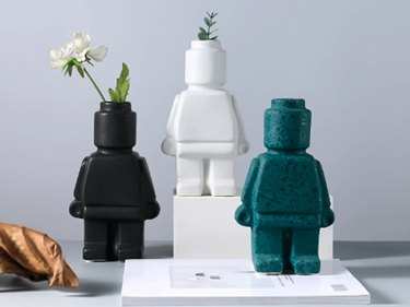 Three vases (in black, white and blue) shaped like large LEGOs. The white and black vases have plants and flowers inside.