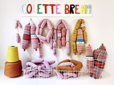 A variety of knitted sausages and salami hanging from wall hooks and strewn in white wire baskets.