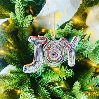 A multicolored ornament made of recycled paper with the word "Joy" hangs from a Christmas tree.