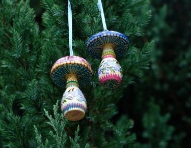 Two hand-painted ornaments shaped like mushrooms dangling from a Christmas tree branch. Both mushrooms are painted in vibrant colors with dots, flowers and stripes.
