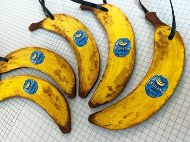 A set of five banana-shaped ornaments cut from wood and painted yellow with darker brown areas. Each banana has a painted-on blue fruit sticker.
