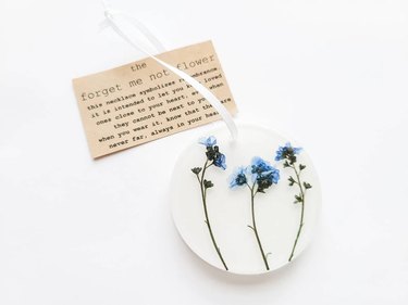 A white circular ornament with three pressed blue forget-me-not flowers inside. The accompanying note is typed on tan paper and explains the sentimental meaning of a forget-me-not.