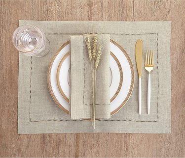 Natural linen placemat from Solino Home, shown in overhead view on a wooden dining table, with a full place setting and matching linen napkn