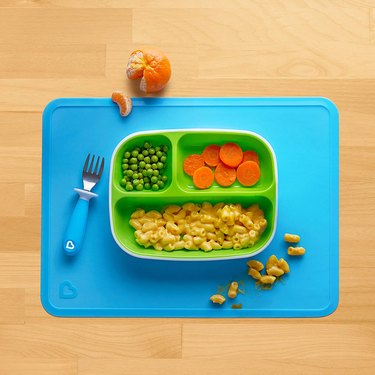 Munchkin silicone placemat in bright blue, shown on a wooden tabletop with a toddler's fork and vegetables in a divided dish