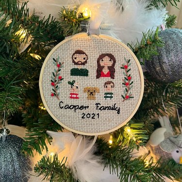 A cross-stitch ornament with two adults, two children, a dog and two decorative branches. The bottom reads, "Cooper Family 2021."