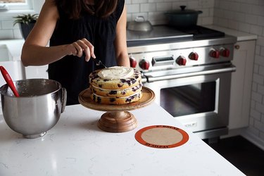 Woman icing a cake on a pedestal, with a round Silpat baking mat in the right foreground and a mixer bowl full of icing on the left