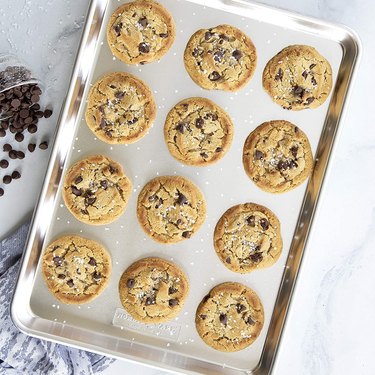 Nordic Ware half-sheet pan, shown in overhead view on a marble countertop, filled with chocolate chip cookies and with a spill of chocolate chips to its left