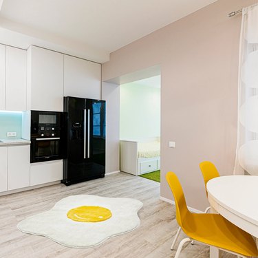 A medium-size accent rug shaped like a white and yellow fried egg sits in the middle of a kitchen, which features tan walls, a white table with yellow chairs and a shiny black refrigerator alongside white cabinets.