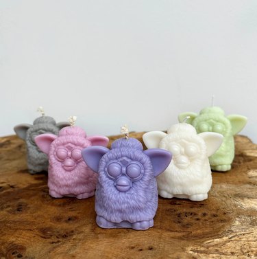 Five pastel-colored candles (gray, pink, purple, white and green) shaped like small gremlin creatures (Furbies) sitting atop a wood table.
