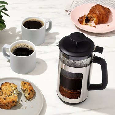 OXO French press on a marble countertop with two cups of coffee and plates of baked goods.