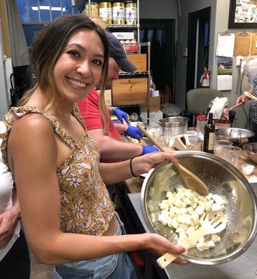 A smiling woman wearing a tank top and light blue jeans stands in a kitchen, mixing croutons in a silver bowl