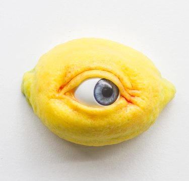 Half a yellow lemon attached to a white wall. The lemon has a fake yet ultra-realistic human eyeball smack dab in the middle.