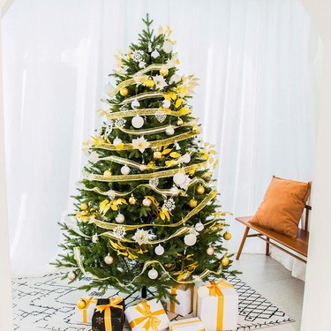 Artificial Christmas Tree with gold ribbon, white ornaments, with yellow and white presents below it.
