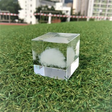 A clear cube with what appears to be a tiny white, wispy cloud in the center sits on a patch of green grass.