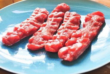 Four pieces of reddish-brown soap shaped like fried bacon sit atop a blue plate.