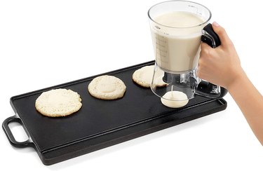 Hand holding an OXO batter dispenser over a griddle, and using it to dispense batter for pancakes