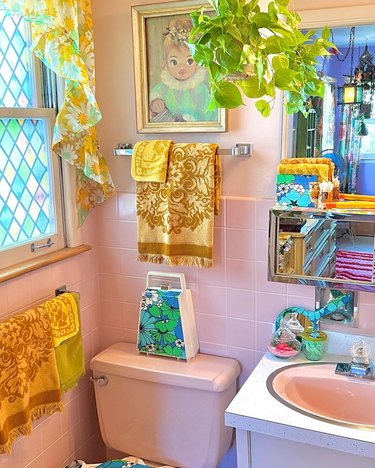 Pink bathroom with yellow hand towels, a stained glass window, greenery and floral curtains