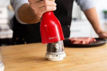 Male cook in black apron, shown pressing a red Finamill spice grinder onto an interchangeable pod in order to season a steak visible in the background