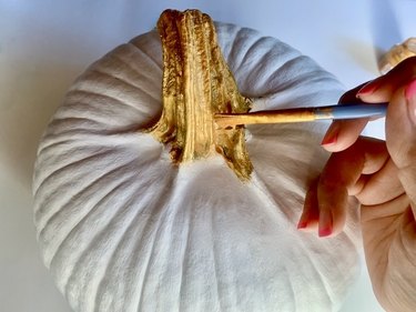 The crafter paints the stem of the white-painted pumpkin with gold paint using a paintbrush