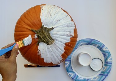 The crafter applies white paint to a medium-size pumpkin