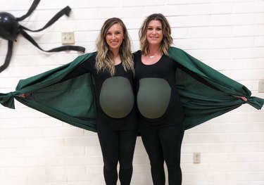Two pregnant women wearing black and green shirts while holding up a green sheet