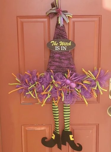 Wreath shaped like purple, green, orange and black witch hat with dangling legs hanging on a red front door