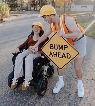 Pregnant person in a wheelchair wearing a hard hat next to a person in an orange vest holding a sign that reads "Bump Ahead"