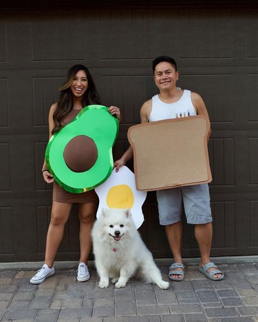 Pregnant woman in an avocado costume standing next to a man in a toast costume and a white dog in a fried egg costume