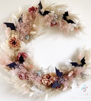 Wreath made with dried flowers and grass, all in neutral tones, and topped with black paper bats