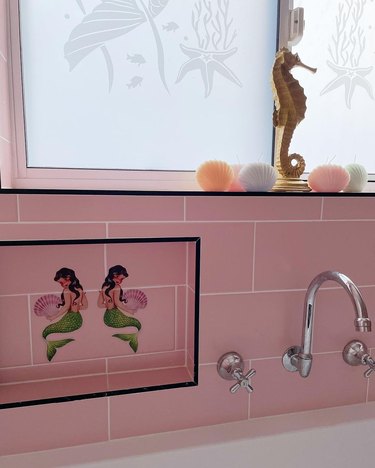 Pink tile bathroom wall next to bathtub with two mermaid decals and a gold seahorse figurine