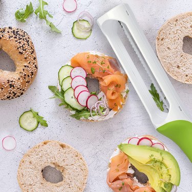 Handheld bagel slicer surrounded by bagels with different toppings on them like lox, avocado and radishes.