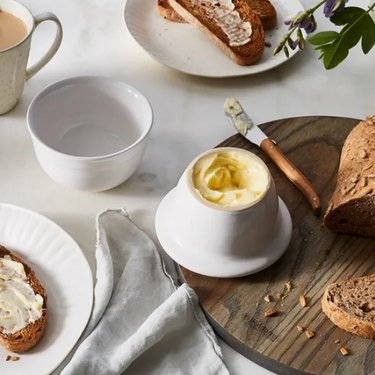 Emile Henry French butter crock, shown on a wooden cutting board with multigrain bread, which in turn rests on a marble countertop with buttered toast and a cup of tea