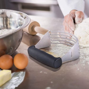 A broad stainless steel pastry blender with a soft black ergonomic handle with finger indentations, next to a hand shaping dough.