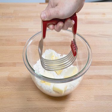 A tall pastry blender with six stainless steel blades, a cleaning tab attached to the blades and a dark red plastic handle