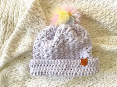 A lavender crochet beanie with a rainbow fur pom pom and leather heart tag on the cuff.