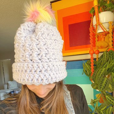 A woman wearing a lavender crochet beanie with a multicolored pom pom on top