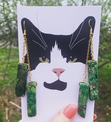 A picture of a black and white cat with actual green stone earrings hanging from the cat's ears