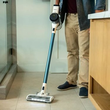 Man using the Tienco S10 cordless vacuum to clean hard surface flooring in a bathroom. The vacuum has a blue shaft and LED lights.