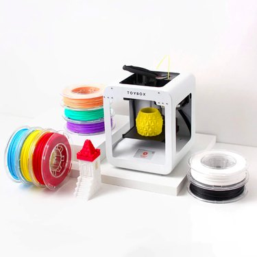 A small white 3D printer box with a yellow toy in the center, surrounded by spools of colorful filament.