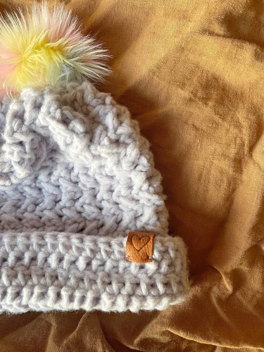 A lavender crochet beanie with a rainbow pom pom and leather hat tag.