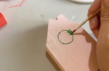 A person painting a green Christmas wreath on a wooden pink house