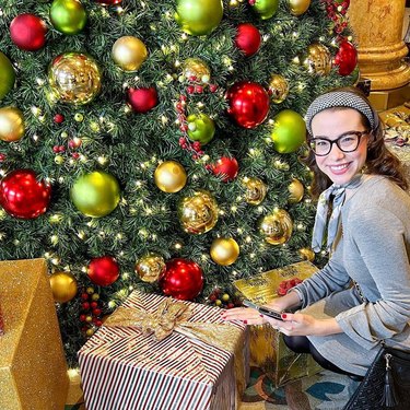 Woman in gray sweater sitting next to a Christmas tree and wrapped holiday gifts