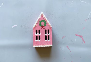 A pink wood house with a Christmas wreath painted in the center and white snow on top.