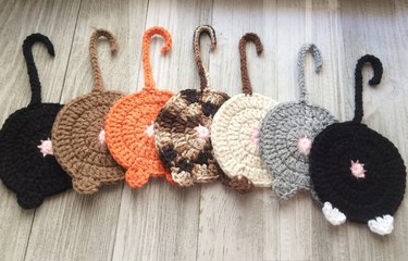 Seven crochet cat butt coasters in various colors (orange, grey, black, brown, etc.) with tails.