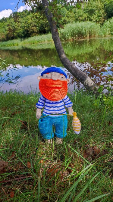 A crocheted fisherman with a bushy orange beard, a striped blue and white shirt, blue pants and a blue beanie. He's holding a yellow and white crocheted fish.