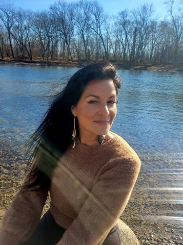 A woman in a fuzzy brown sweater and dangly earrings sits in front of a river