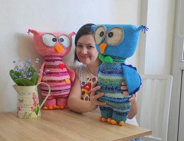 A woman sits between two crocheted stuffed owls, one in pink and orange and the other in blue and green.